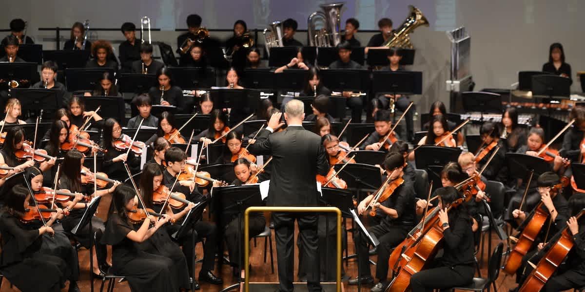 クアラルンプール・インターナショナルスクール: A conductor leads a large orchestra of young musicians playing various instruments.